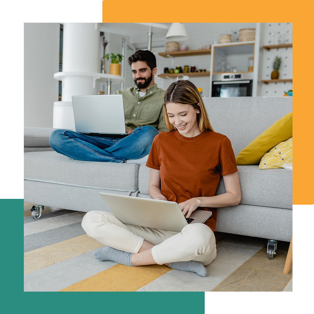 Two people, one sitting on the couch and the other sitting on the ground on their laptop.