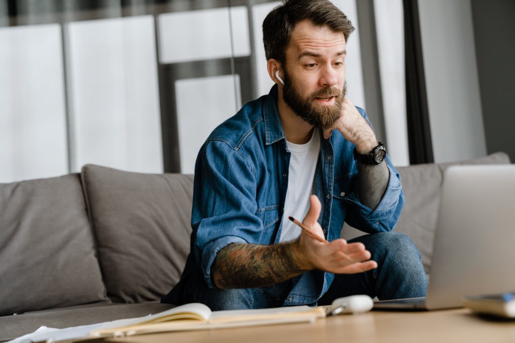 Bearded man gesturing and working with laptop while sitting on sofa.